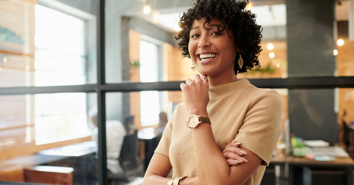 The image shows a young woman with dark curly hair and a warm smile, standing in front of a glass wall, showcasing a modern office setting. She is wearing a beige sweater and a gold watch, with her arms crossed, suggesting a confident and professional demeanor. The background is blurred but hints at a vibrant workspace. The overall impression is one of success and positive energy.